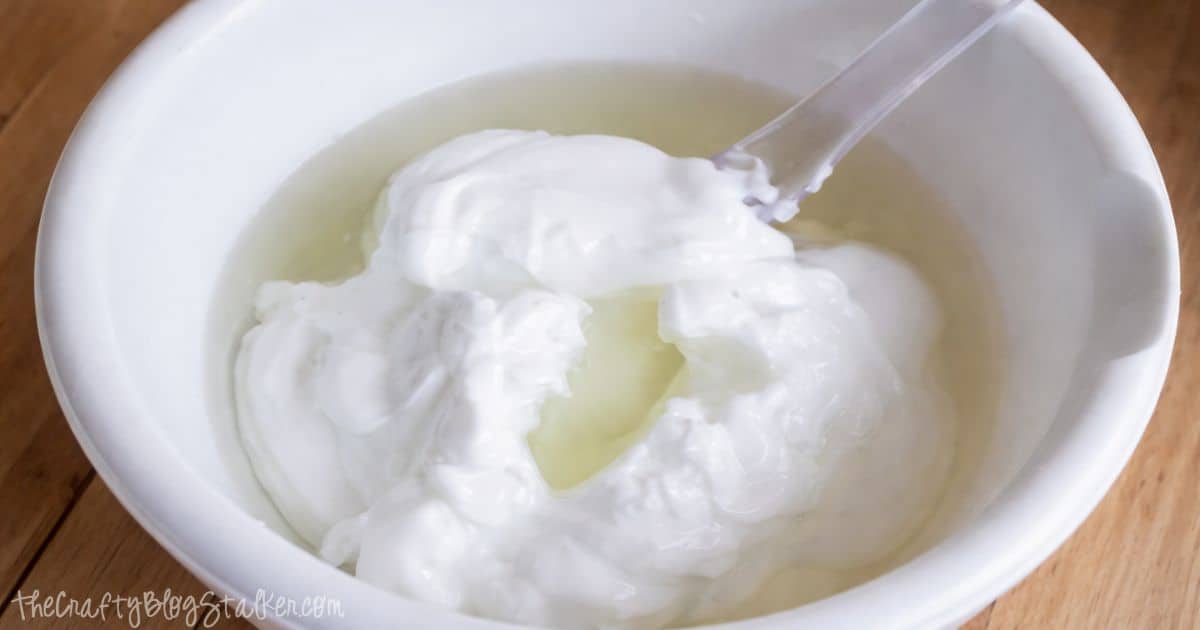 White bowl with thick lotion mixture and oil being combined, with a spoon partially stirring the ingredients together.
