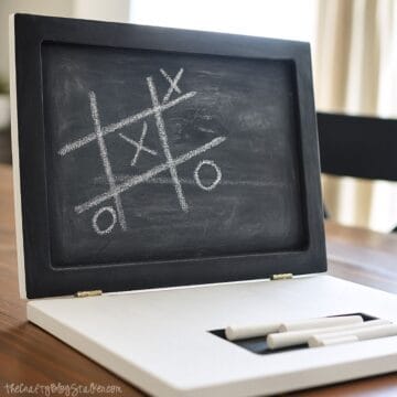 A wooden chalkboard laptop for toddlers with a tic-tac-toe game drawn on the screen and chalk pieces stored in a recessed slot on the base.