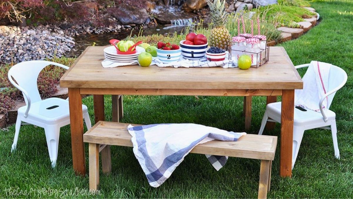 DIY kids outdoor table with bench seating, white chairs, and colorful picnic snacks set up on a grassy backyard lawn.