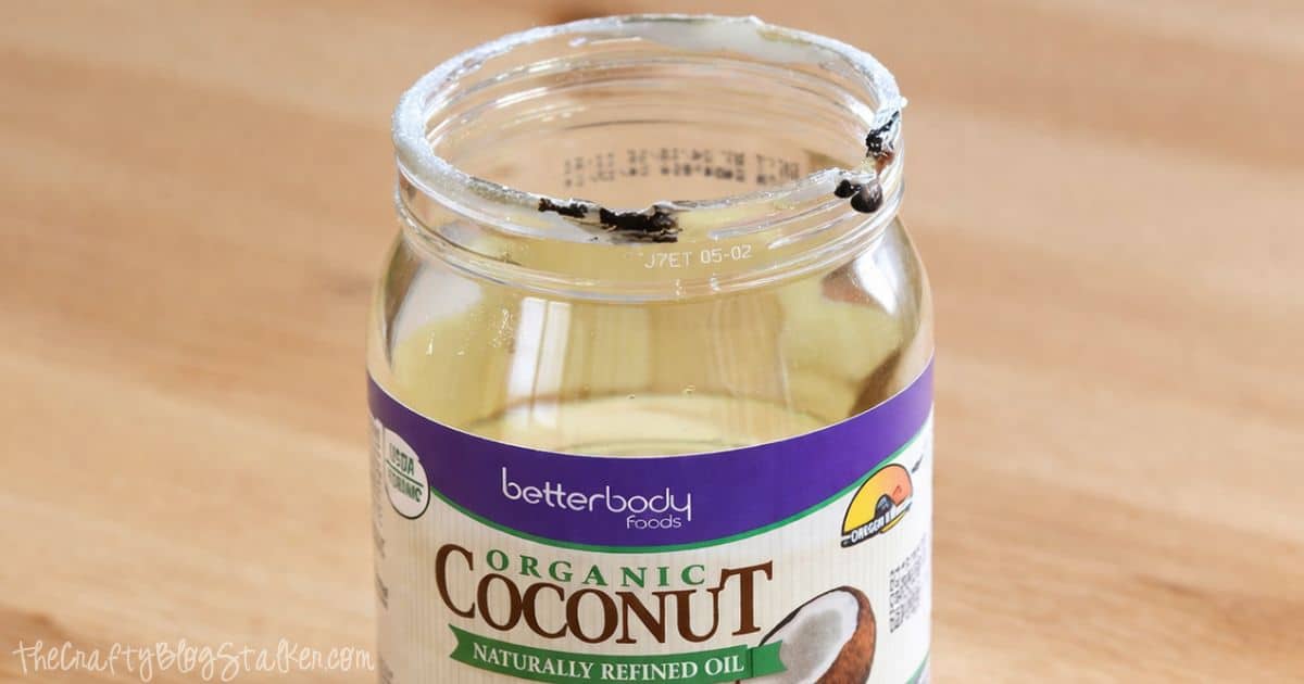Open jar of melted coconut oil with a label on the front, sitting on a wooden surface with oil residue around the rim.