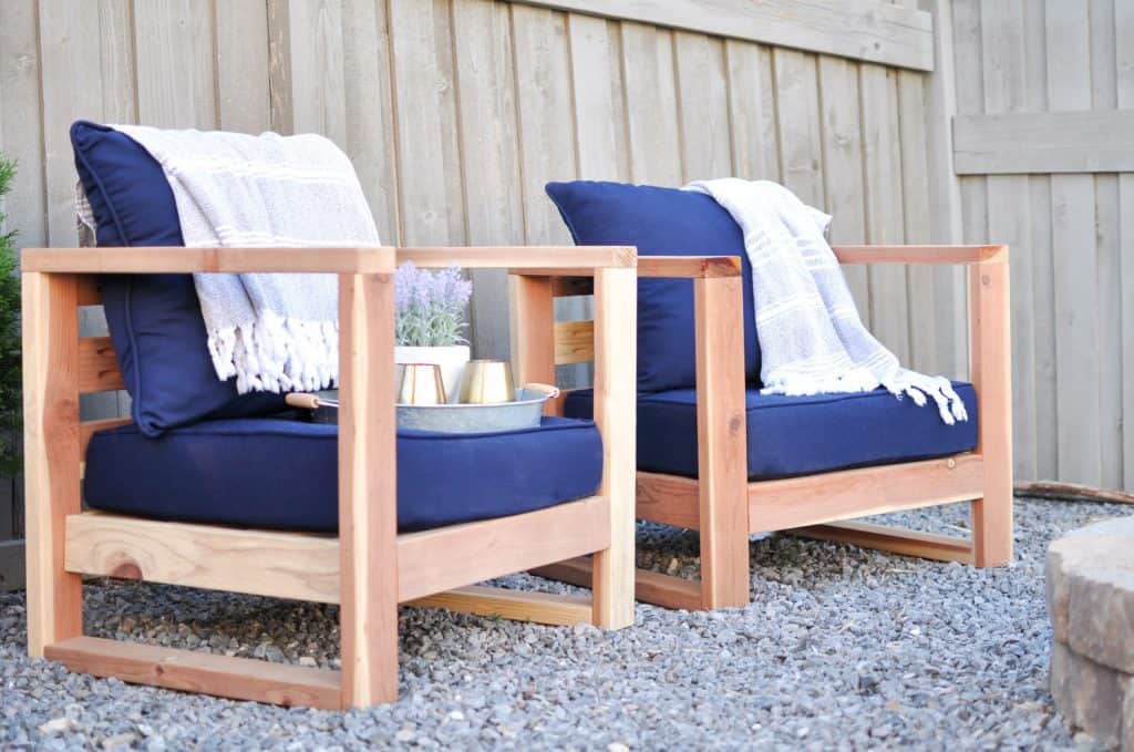 Two modern wooden outdoor chairs with navy blue cushions and white throw blankets, placed on gravel near a wooden fence with a small tray of drinks between them.