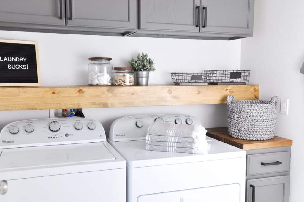 Modern farmhouse laundry room with gray cabinets, a wooden shelf over Whirlpool washer and dryer, labeled board, storage jars, baskets, and a crocheted bin.