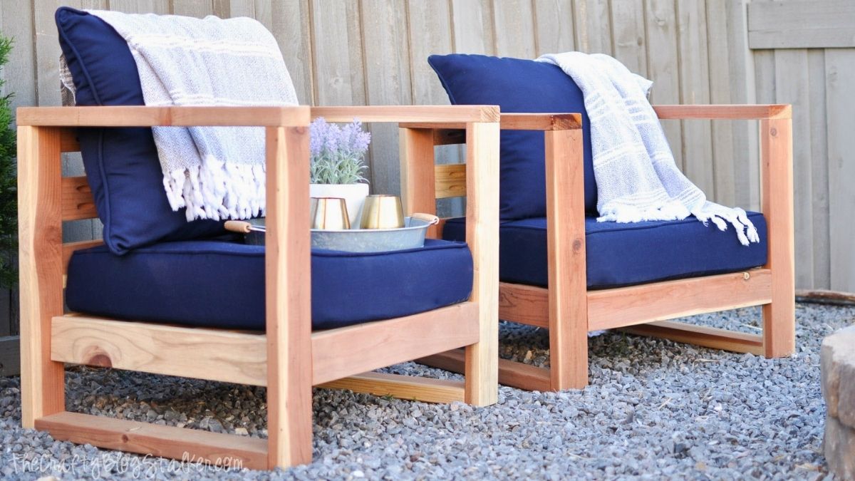 Two DIY wooden outdoor chairs with navy blue cushions and white throws on a gravel patio beside a wooden fence.