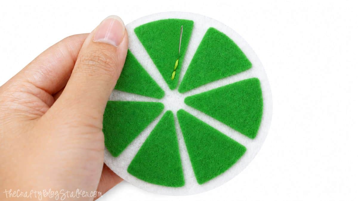 Hand stitching green felt citrus segments onto a white felt coaster base to create a DIY lime slice coaster.