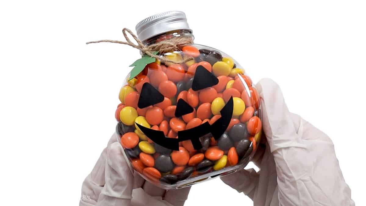 Clear jack-o'-lantern candy jar filled with orange, yellow, and brown candies, decorated with a black vinyl face, twine bow, and green leaf.