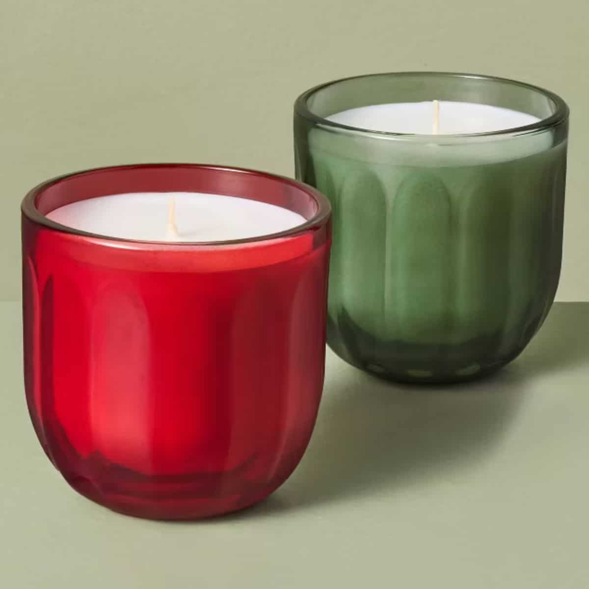 Two unlit candles with white wax in red and green glass jars featuring a ribbed design, displayed against a sage green background.