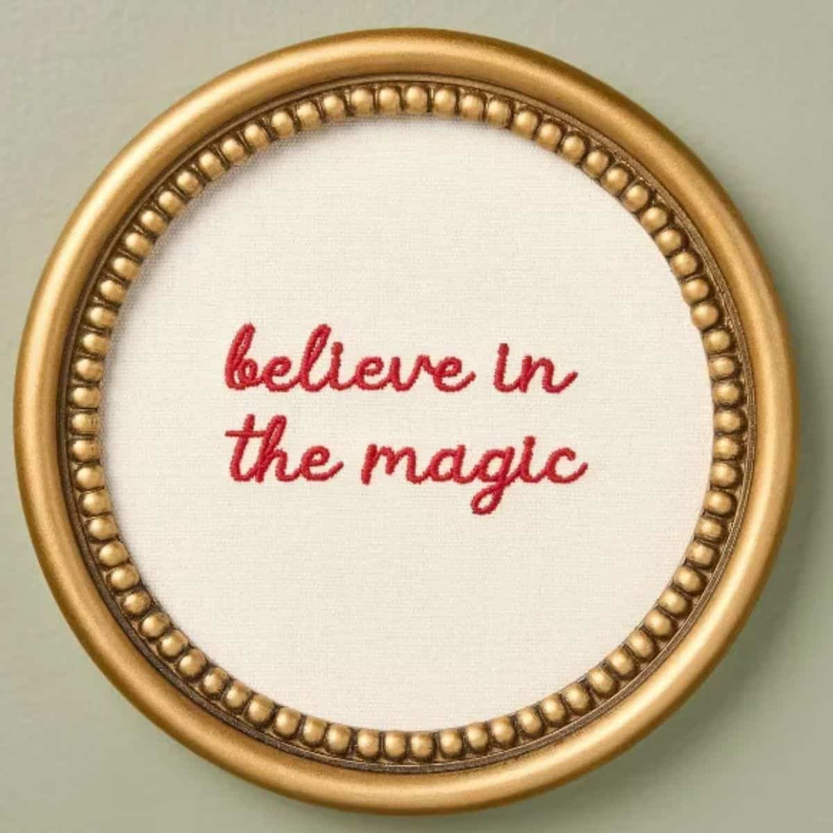 Round gold beaded frame with white fabric embroidered with the words "believe in the magic" in red cursive lettering.