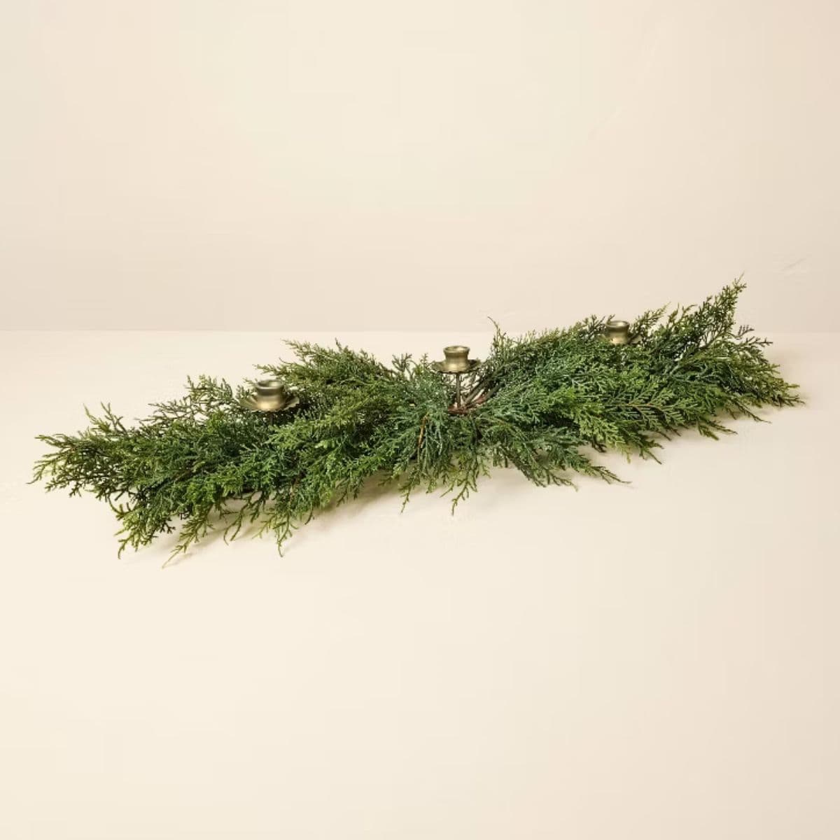 Christmas greenery centerpiece with lush evergreen branches and three brass candle holders arranged on a cream-colored surface.