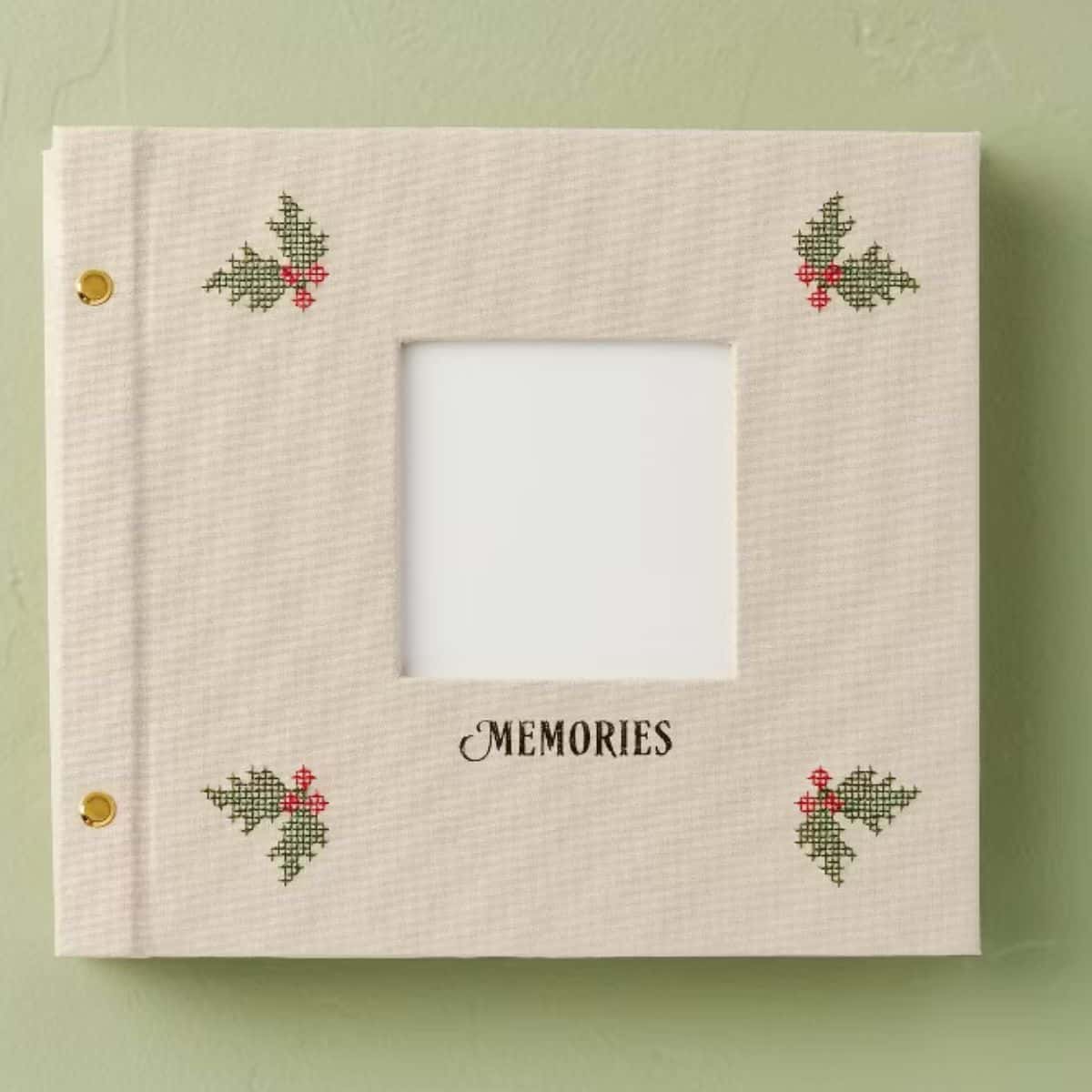 Cream fabric memory book with cross stitch holly leaves and red berries in each corner, a square photo opening, and the word "Memories" on the cover.