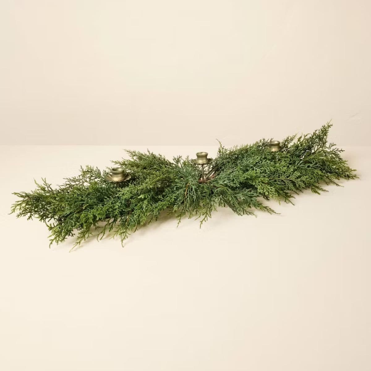 Greenery table centerpiece with three brass candle holders arranged among lifelike evergreen branches for a festive holiday display.