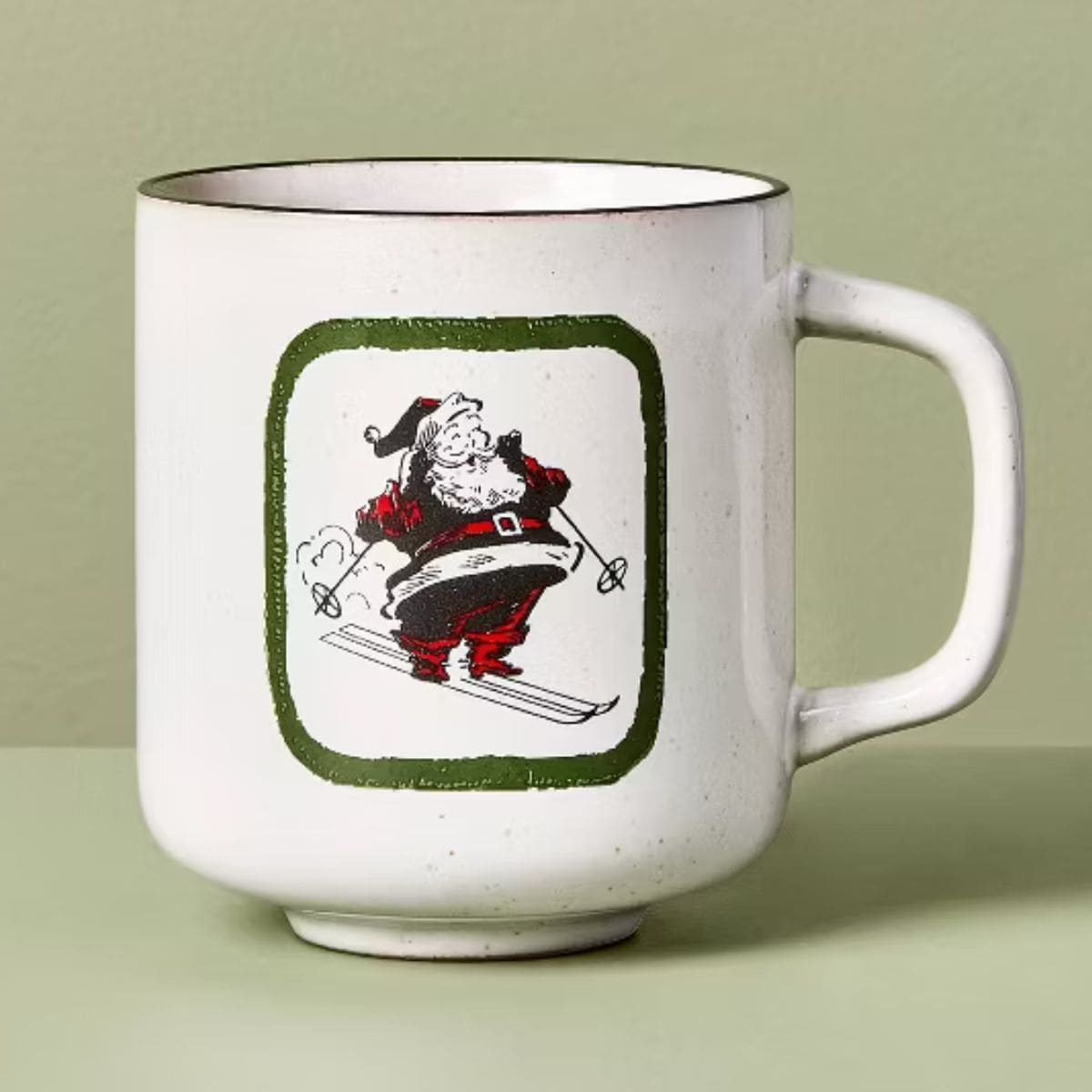 White ceramic holiday mug with a green border and a vintage-style illustration of Santa Claus skiing, dressed in his red suit and hat.