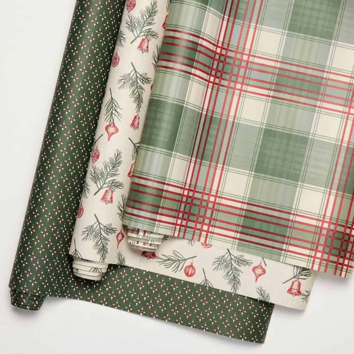 Three rolls of Christmas wrapping paper in green and red designs, including plaid, pine sprigs with ornaments, and a small holly pattern.