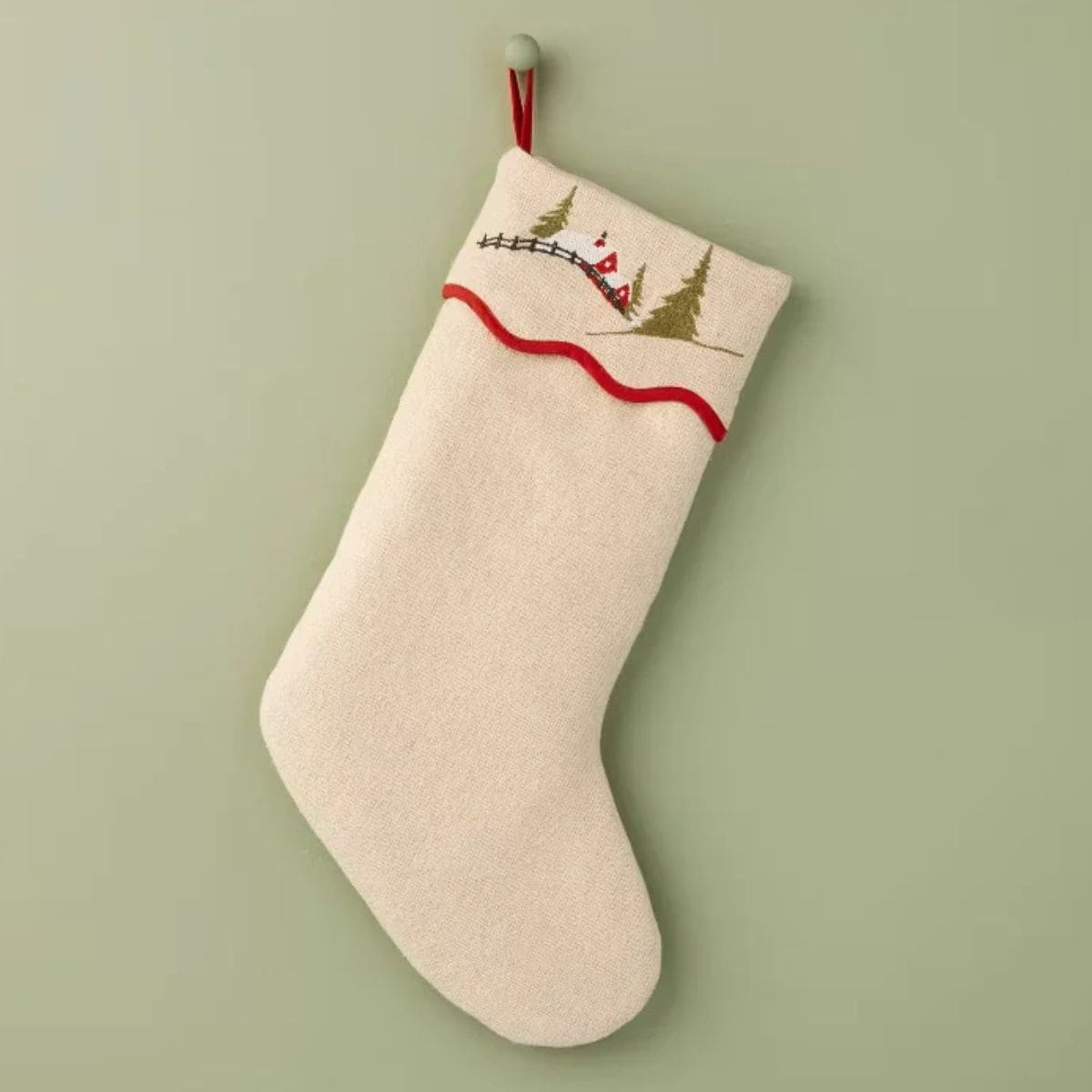 Cream-colored Christmas stocking with embroidered evergreen trees, red accents, and wavy red trim hanging against a sage green wall.