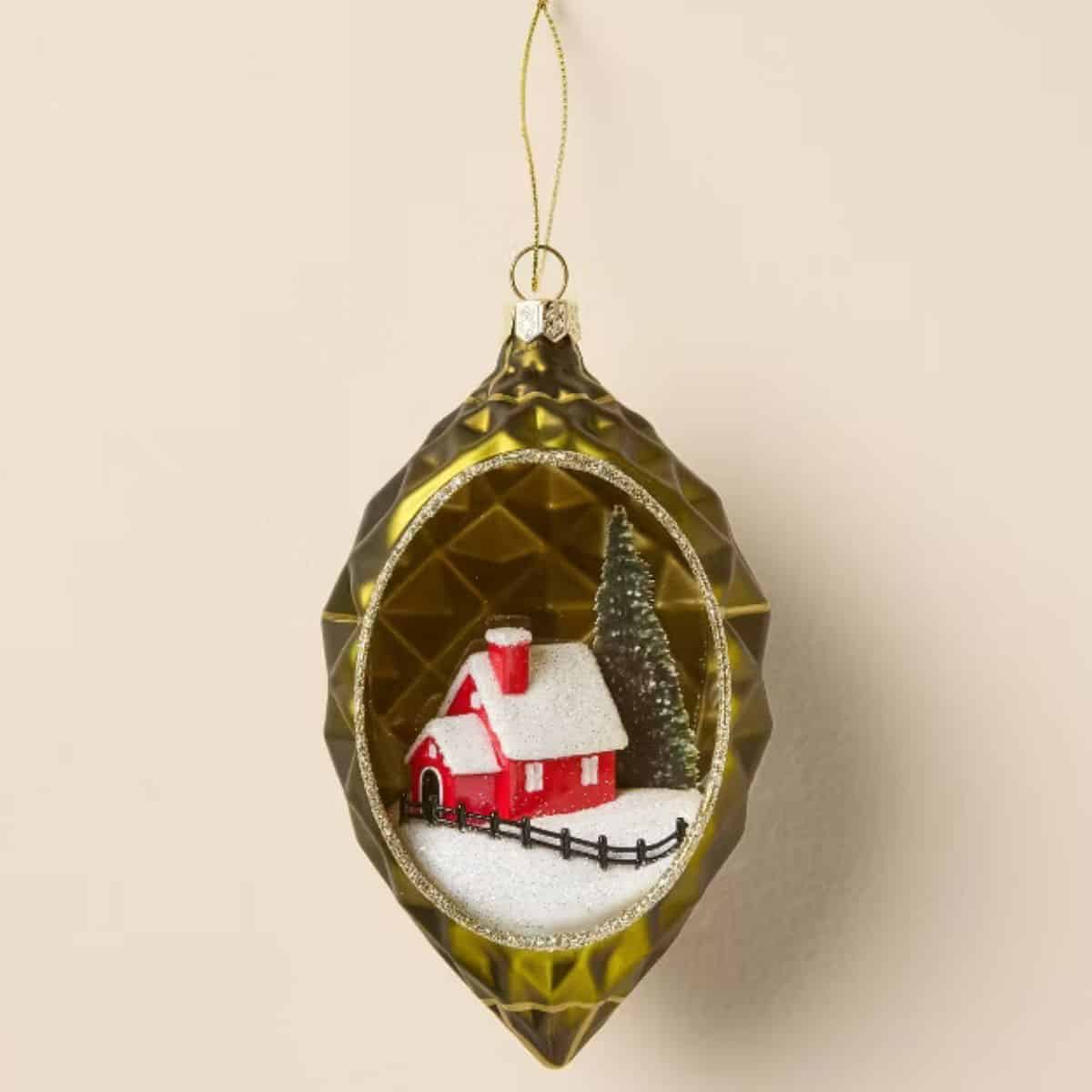 Gold glass Christmas ornament featuring a snowy red house, evergreen tree, and black fence inside a glitter-trimmed oval opening.