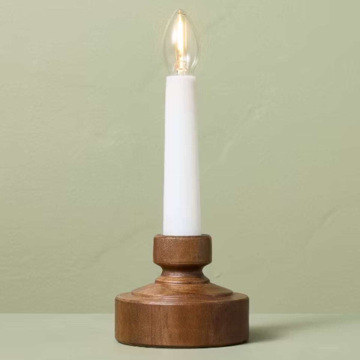 White flameless taper candle with a glowing bulb displayed in a brown wooden candle holder against a sage green background.