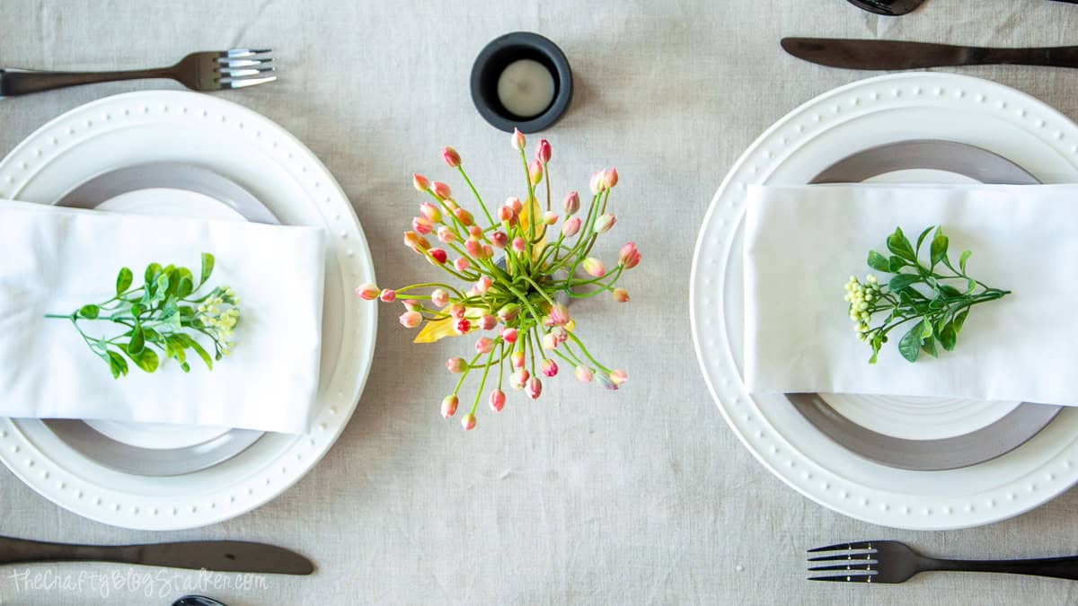 Overhead view of a summer tablescape with white plates, folded napkins topped with greenery sprigs, black flatware, and a floral centerpiece on linen.
