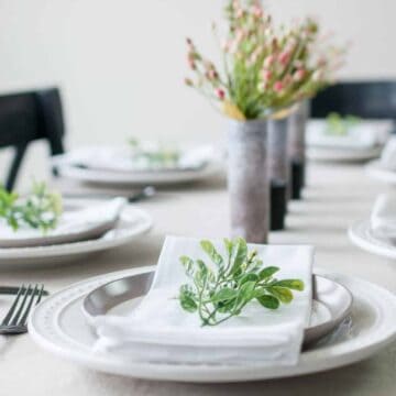 Simple summer tablescape with layered white plates, folded napkins, greenery accents, and floral centerpieces on a neutral linen-covered dining table.