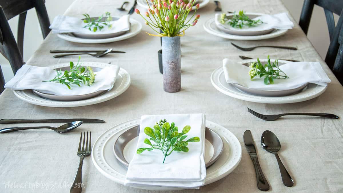 Summer tablescape with white plates, black flatware, greenery-topped napkins, and floral centerpieces arranged on a neutral linen-covered dining table.