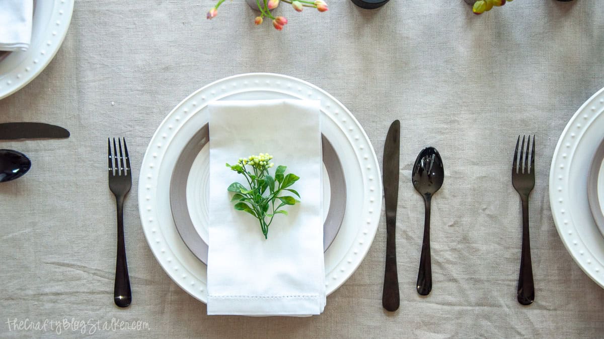 Summer table place setting with layered white plates, a folded white napkin, black flatware, and a small greenery sprig on a neutral linen tablecloth.