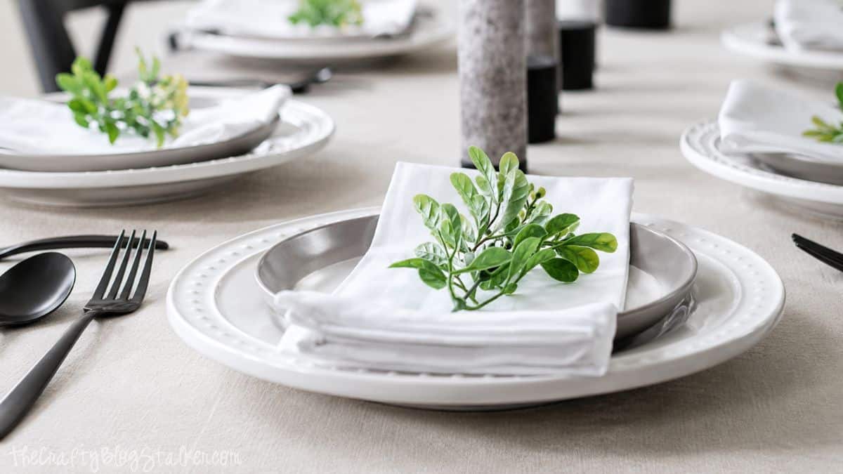 Close-up of a summer table setting with layered white plates, a folded white napkin, greenery accent, and black flatware on a linen tablecloth.