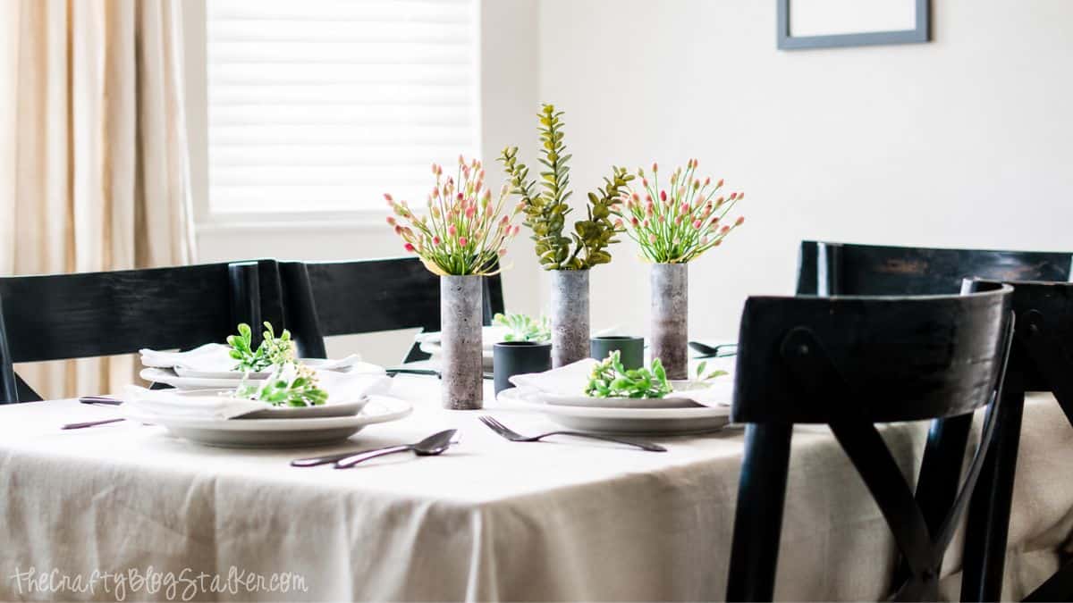 Side view of a summer dining table with white place settings, greenery accents, black chairs, and floral centerpieces on a neutral tablecloth.