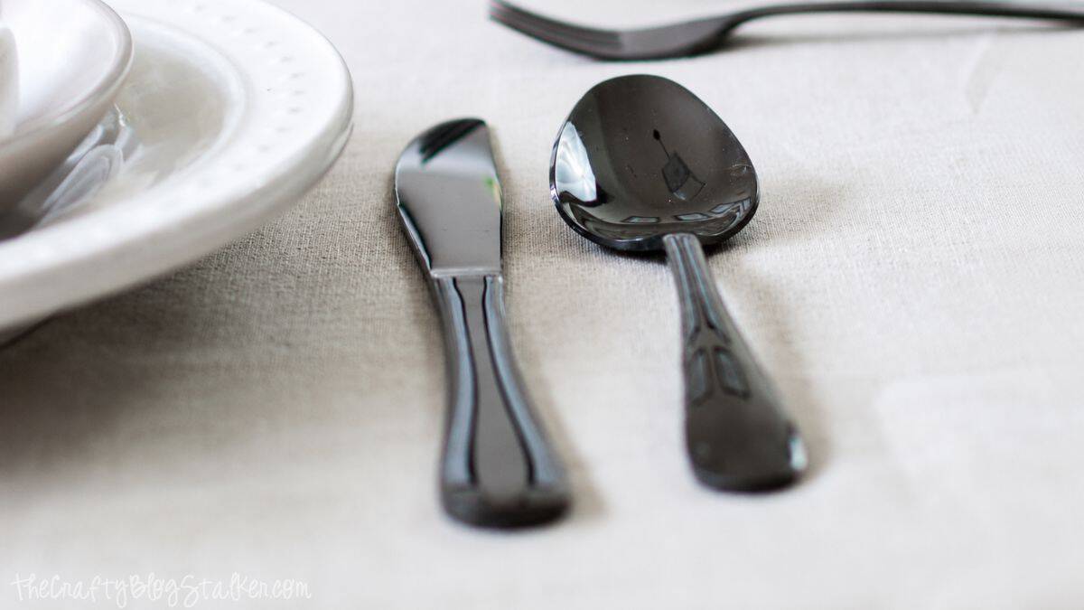 Close-up of black spoon and knife beside a white plate on a neutral linen tablecloth, highlighting a simple and modern summer table setting.