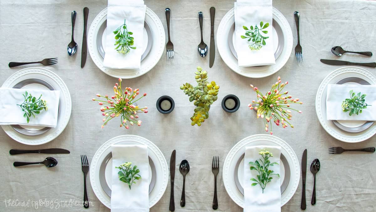 Overhead view of a summer dining table with six place settings, white plates, black flatware, greenery accents, candles, and floral centerpieces.