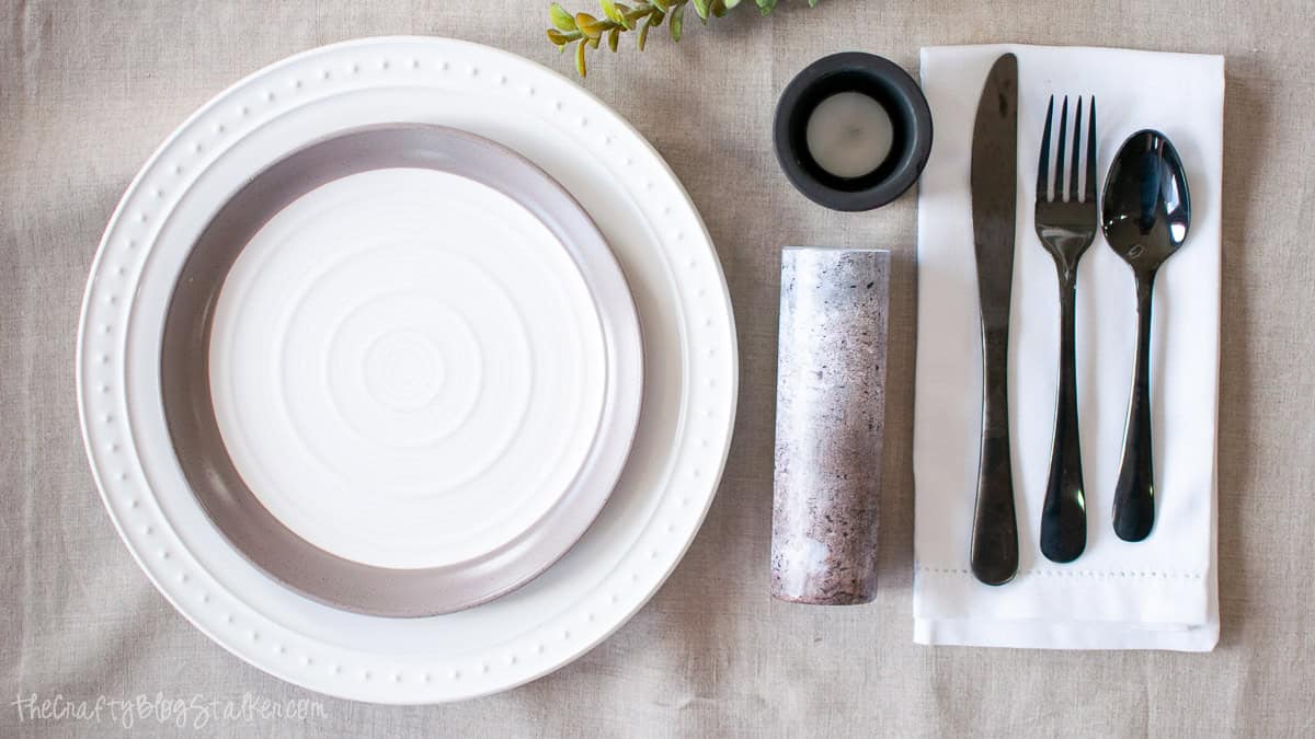 Flat lay table setting with white dinnerware, black silverware on a folded napkin, candle holder, cork, and greenery on a neutral linen tablecloth.