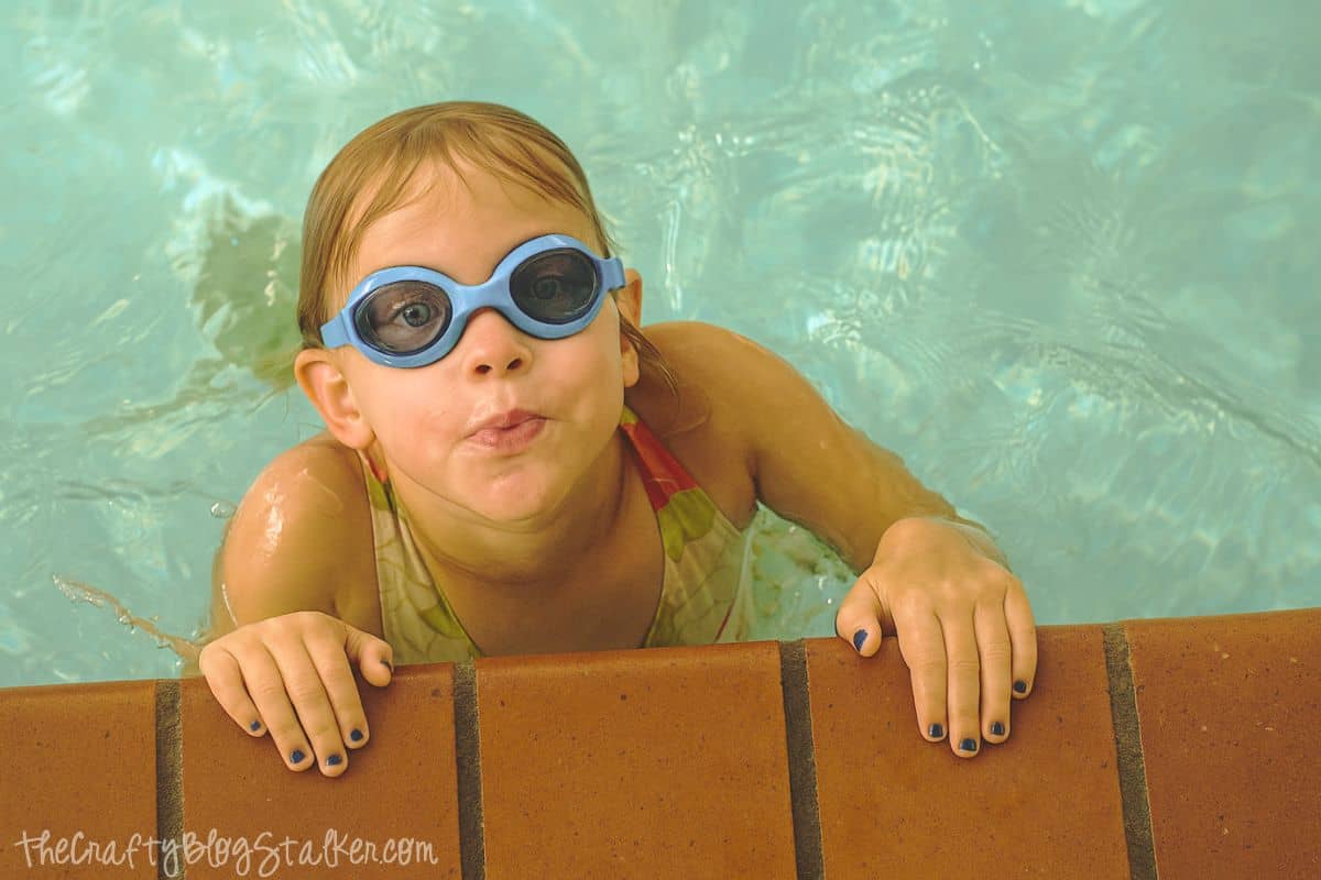 Young girl wearing blue swim goggles playfully clings to the edge of a swimming pool, smiling with her nails painted blue.