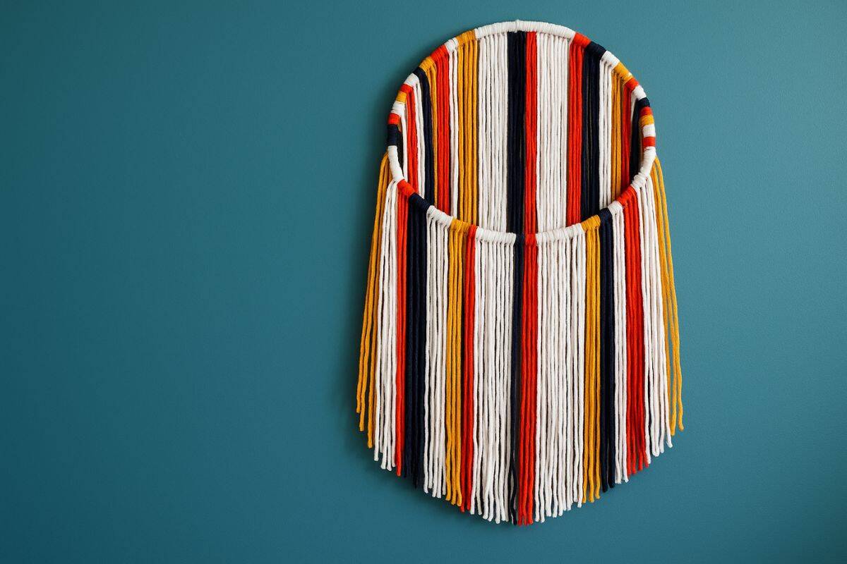 Colorful circular yarn wall hanging with vertical strands in white, black, red, and yellow, draped from a round frame against a teal wall background.