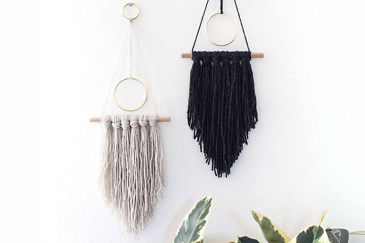 Two modern yarn wall hangings in beige and black, each suspended from a wooden dowel and gold ring, displayed against a white wall above green houseplants.