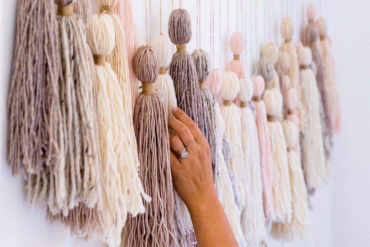 Hand touching a wall display of large yarn tassels in soft neutral tones like cream, taupe, and blush, tied at the top and hanging in a row for a cozy decor look.