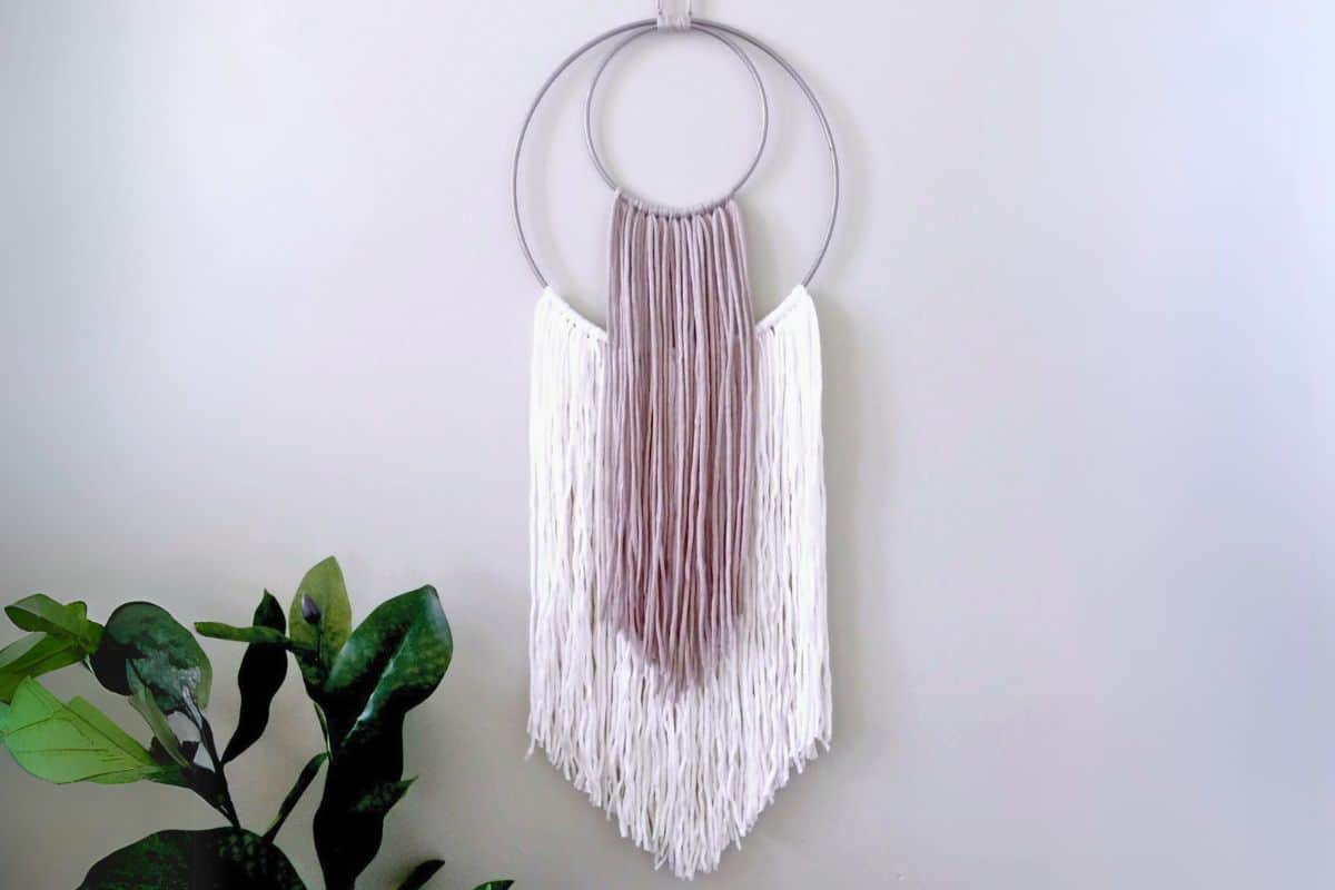 Modern boho yarn wall hanging with layered metal hoops and neutral-toned yarn in taupe and white, styled against a pale wall above leafy green houseplants.