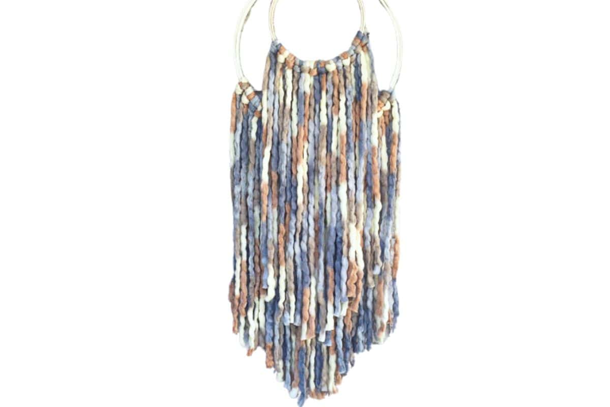 Wall hanging made with chunky multicolored yarn in blue, tan, and cream tones, looped through two metal rings in a V shape for a cozy, boho-style decor piece.