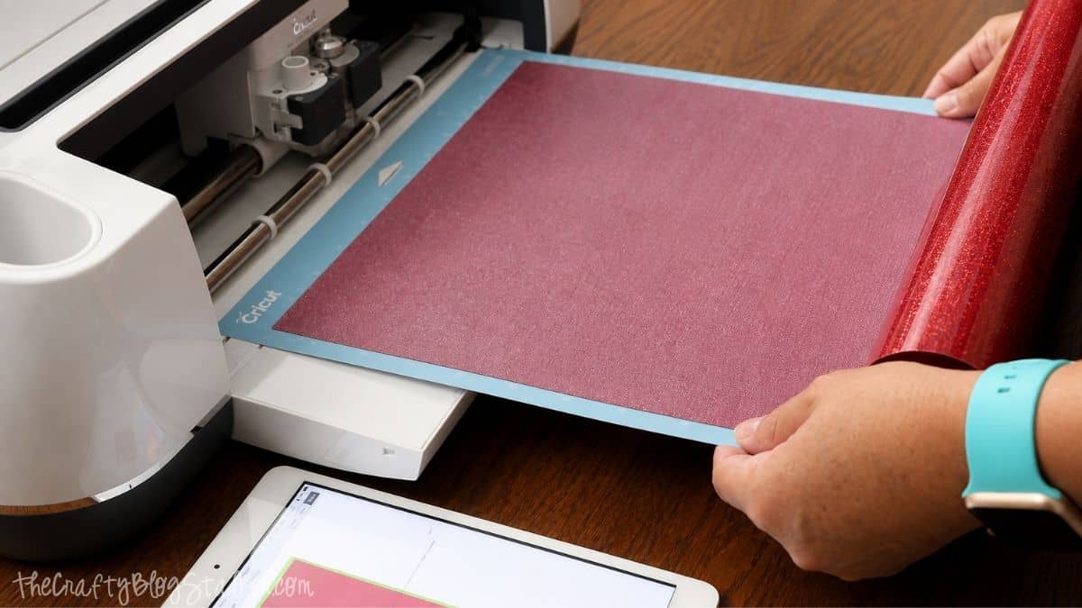 Hands loading a Cricut cutting mat with red glitter iron-on vinyl into a Cricut machine for a Halloween T-shirt design.