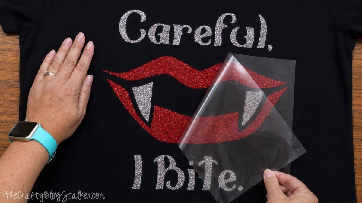 Hands peeling the clear carrier sheet from red glitter vampire lips layered over silver glitter fangs on a black Halloween T-shirt.