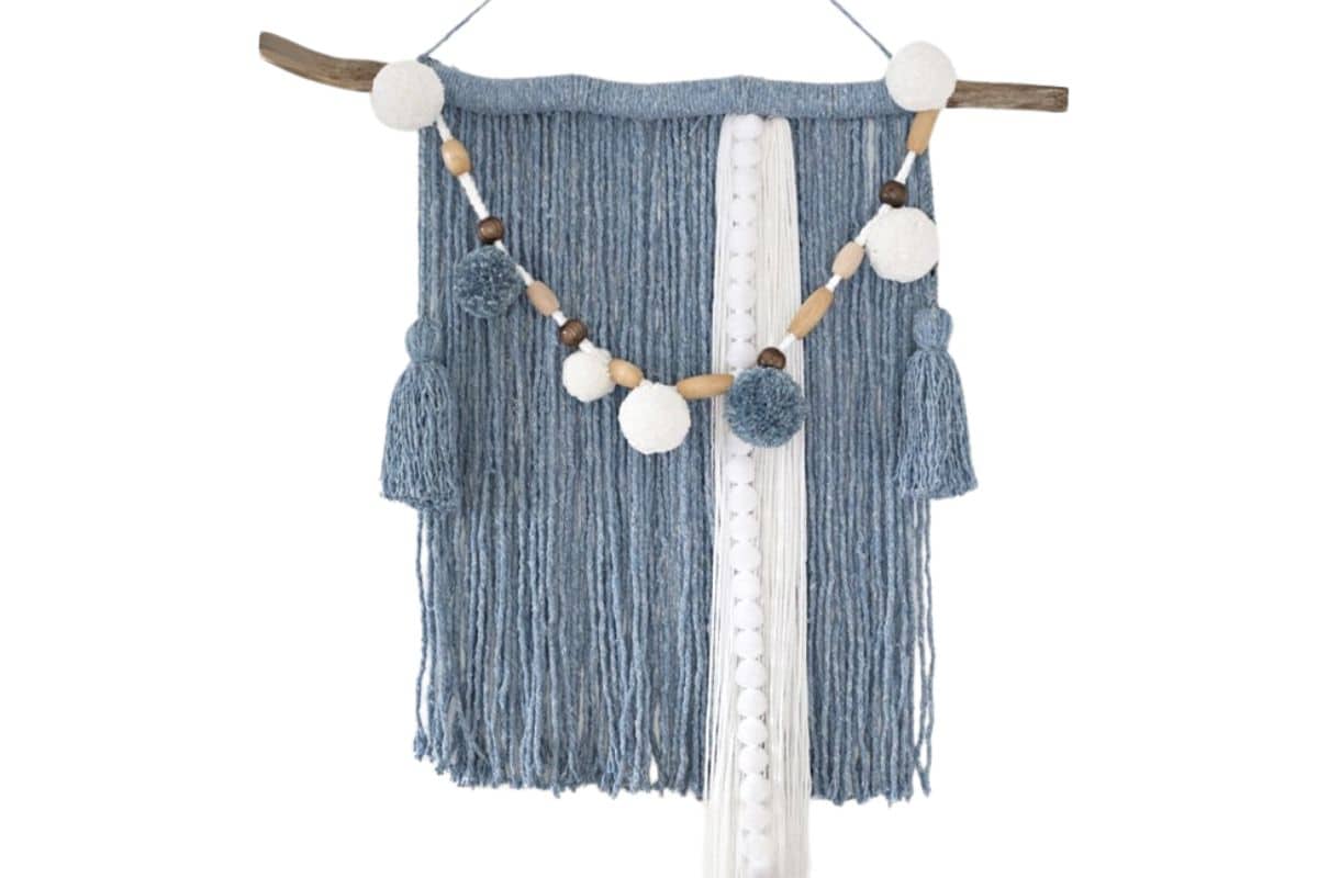 Blue and white yarn wall hanging made from a mop head, decorated with tassels and a garland of pom-poms and wooden beads, hanging from a rustic branch.