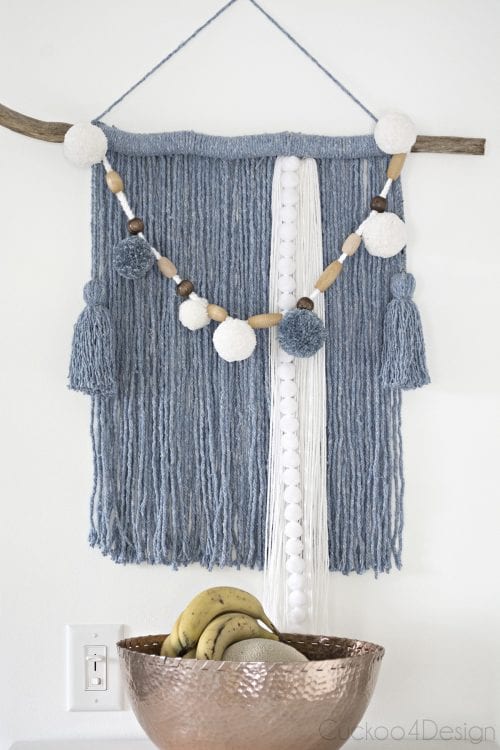 Dusty blue and white yarn wall hanging with a pom pom sash.