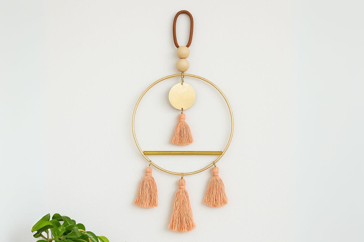Scandinavian-inspired DIY wall art made with a gold hoop, wooden beads, peach yarn tassels, and a brass disc, hanging against a light-colored wall.