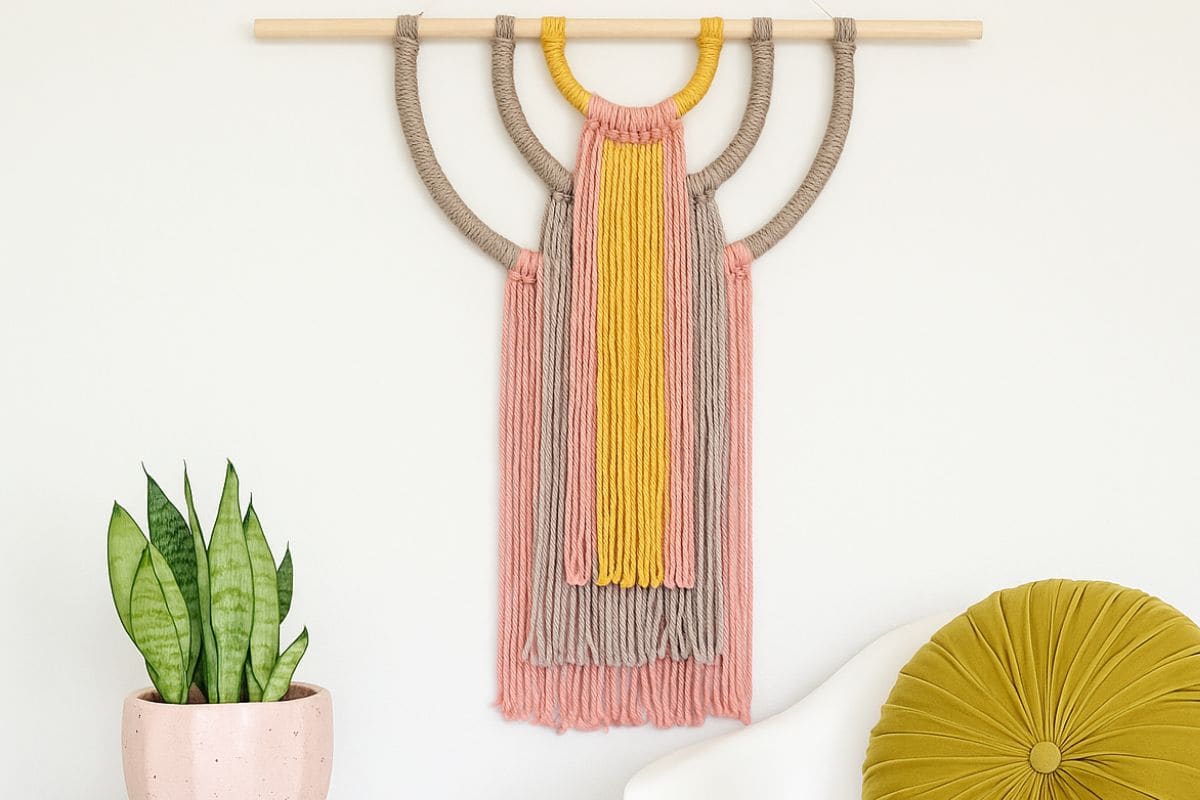 Colorful yarn wall hanging with pink, tan, and yellow strands arranged in layered arches, displayed above a modern white chair and a potted snake plant.