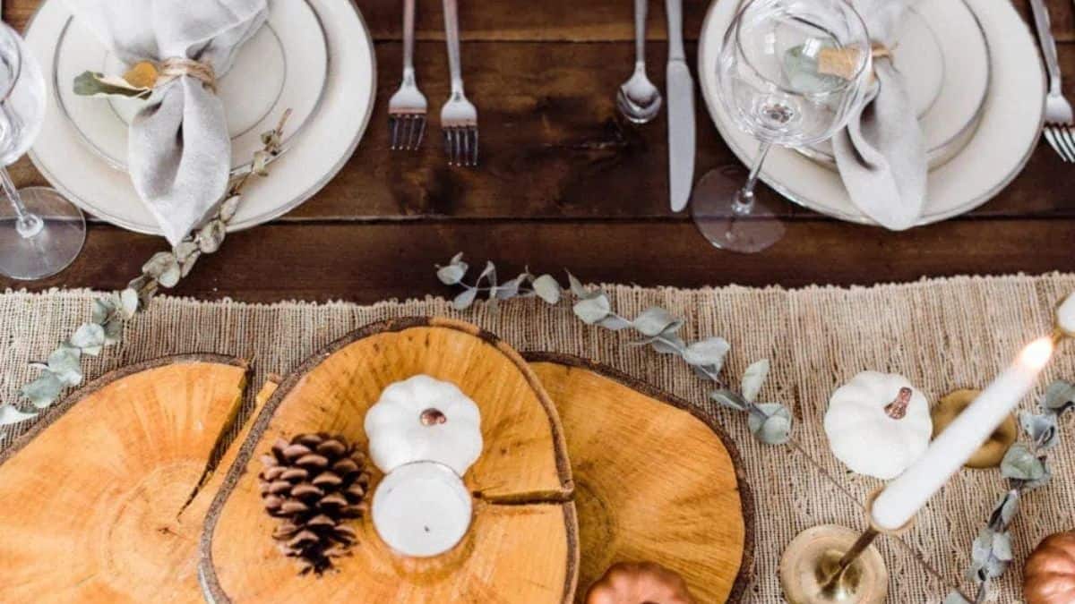 Rustic Thanksgiving table with wood slice centerpieces, white pumpkins, pinecones, candles, and neutral place settings on a burlap runner.