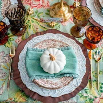 Elegant Thanksgiving place setting with layered plates, a teal napkin, white pumpkin decor, and amber glassware on a floral tablecloth.