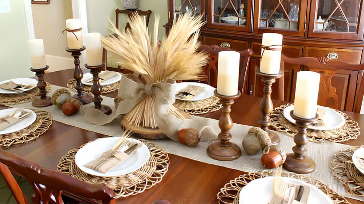 Elegant fall dining table with a wheat bundle centerpiece, pillar candles, woven placemats, and neutral place settings on a wooden table.