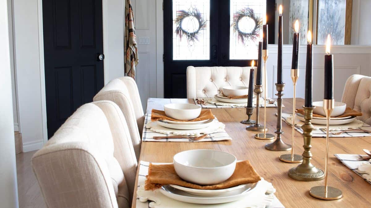 Elegant farmhouse Thanksgiving table with neutral place settings, black candles in brass holders, and soft upholstered chairs in a cozy dining room.