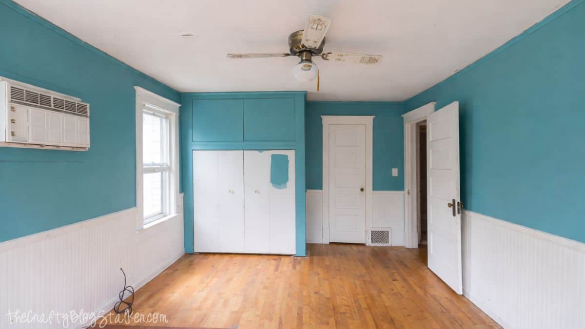 Empty bedroom with hardwood floors, built-in closet cabinets, window air conditioner, ceiling fan, paneled walls, and open doors showing the room before decorating.