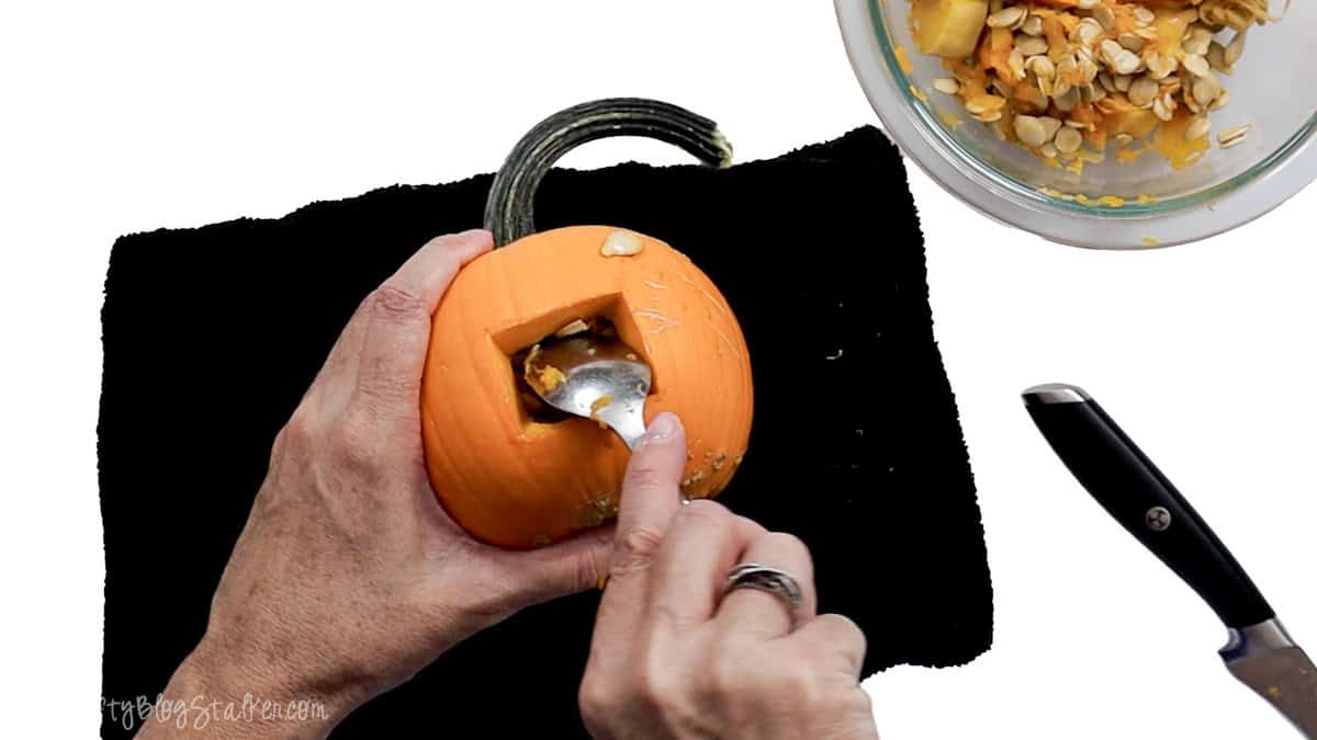 Scooping seeds and pulp from a carved mouth opening in a small orange pumpkin to prepare it for vampire fangs.