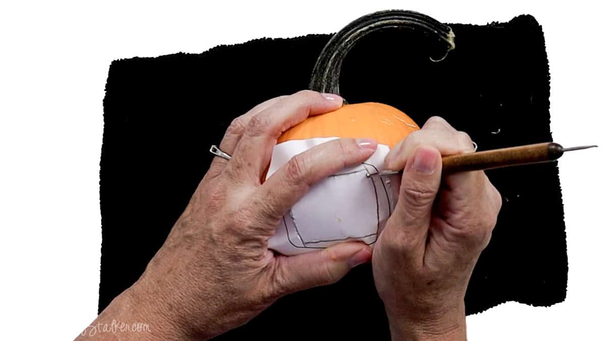 Tracing a vampire mouth template onto a small orange pumpkin with a pointed carving tool before cutting the opening.