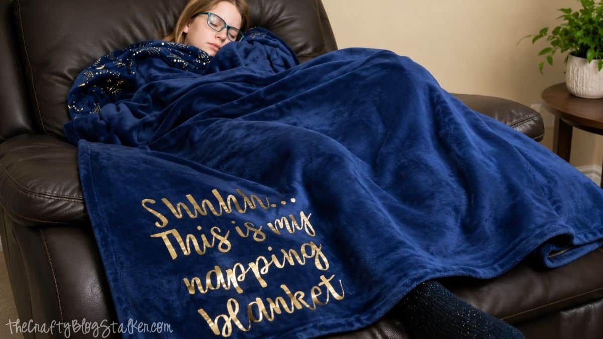 A girl sleeps on a couch wrapped in a plush blue blanket with gold lettering that reads, "Shhhh... This is my napping blanket.