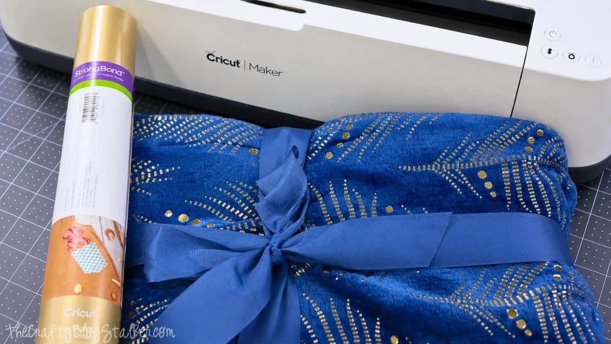 Alt text: A folded plush blue blanket with gold accents tied with a satin ribbon, placed next to a roll of Cricut StrongBond iron-on vinyl, with a Cricut Maker machine in the background.