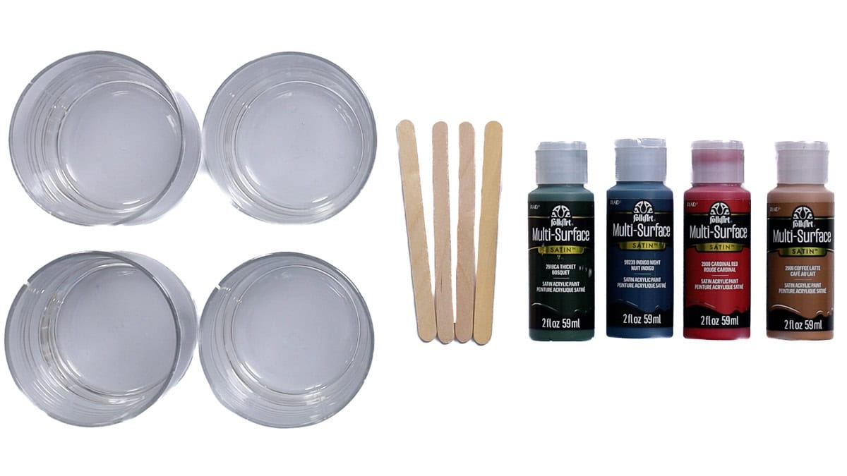 Supplies for a paint glass project including four clear glasses, wooden craft sticks, and four bottles of multi-surface acrylic paint in assorted colors.